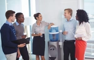 A diverse groups of five people are talking around a water cooler.
